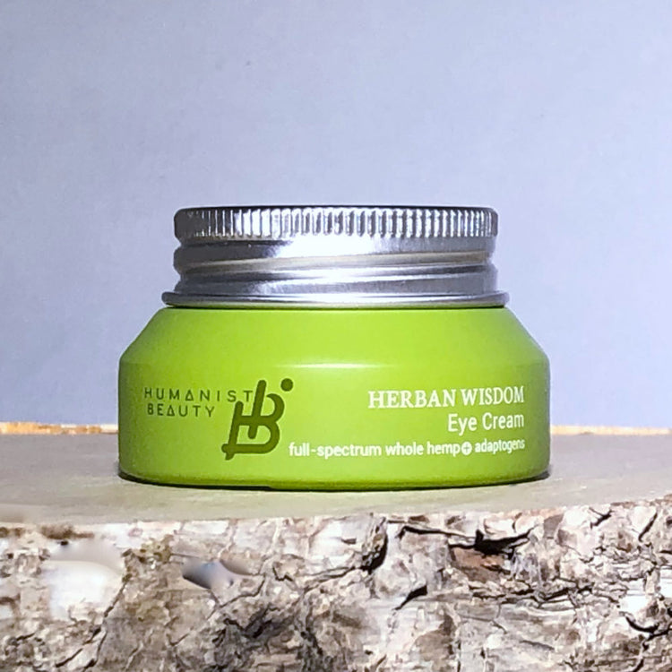 Green jar of Herban Wisdom Eye Cream on a wooden surface with a light blue background